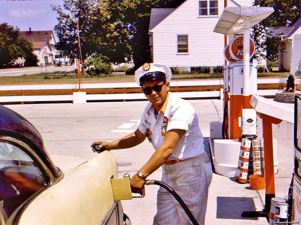A gas station attendant in a white uniform and cap fills up a classic car at an old-fashioned gas pump on a sunny day, with houses and trees visible in the background.