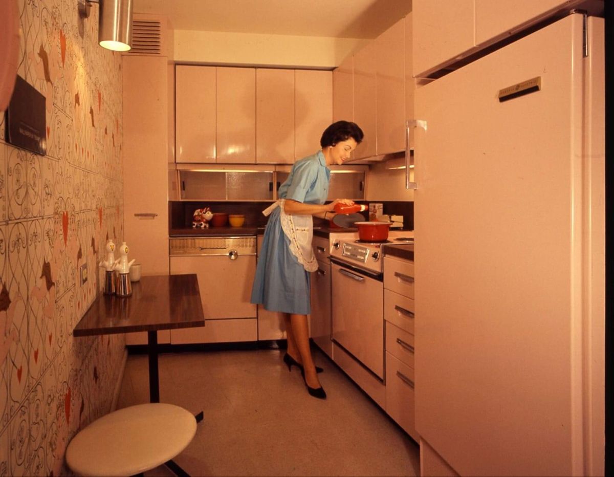 A woman in a blue dress and apron cooks at a stove in a retro pink kitchen with matching cabinets, a refrigerator, patterned wallpaper, and a small dining area.