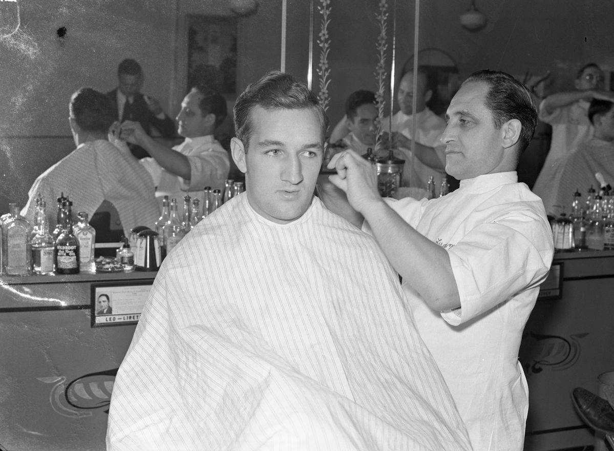 A barber in a white coat gives a haircut to a man in a striped cape. Bottles and tools line the counter, and mirrors reflect other customers and barbers in the busy barbershop.