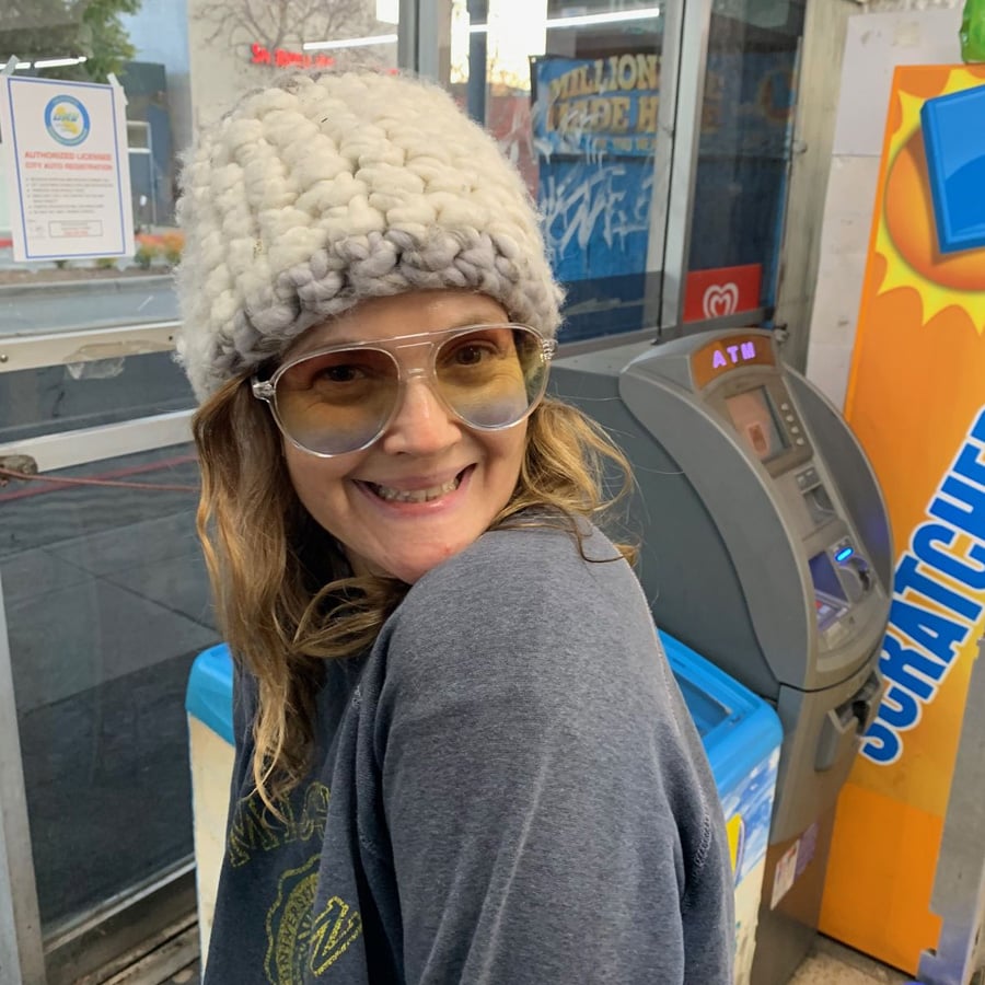 A smiling woman wearing a chunky knit beanie, aviator glasses, and a sweater stands indoors near an ATM and a scratch-off lottery machine.