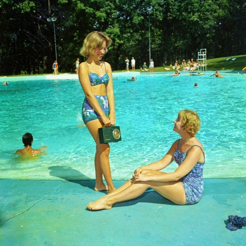 Two women in vintage swimsuits relax by a pool. One stands holding a radio and smiles, while the other sits on the poolside looking up. Trees and other swimmers are visible in the background.