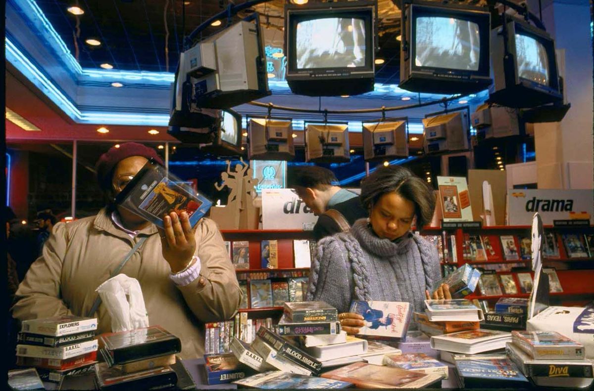 Two people browse VHS tapes in a busy video store with shelves of tapes and several TVs overhead playing movies. The store has bright lighting and a neon ceiling.