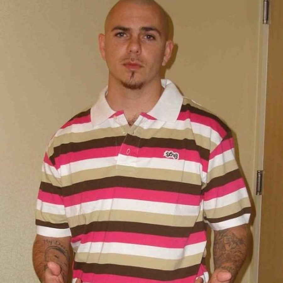 A man with a shaved head and goatee stands indoors, wearing a pink, white, and brown striped polo shirt. He has tattoos on his forearms and is posing with his hands slightly raised.