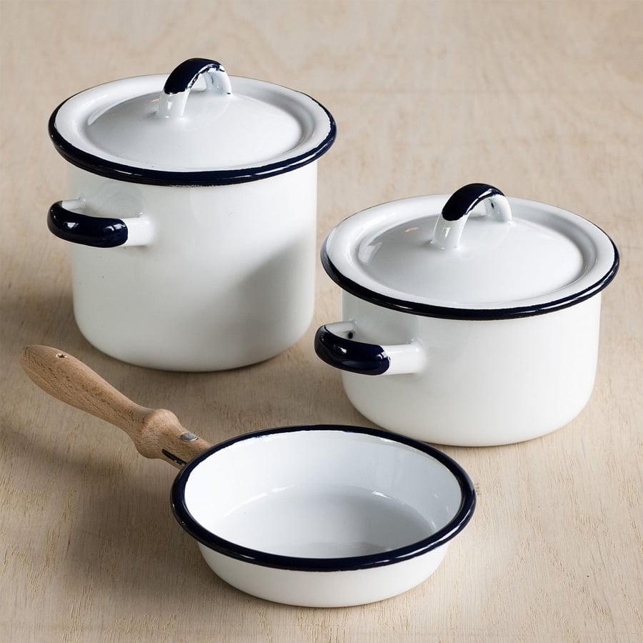 Three white enamel pots with dark rims and lids are on a light wooden surface. Two pots have lids and handles, while the third item is a small pan with a wooden handle and no lid.