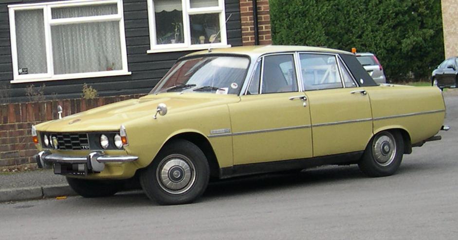 A beige vintage sedan car is parked beside a sidewalk in a residential area, with brick houses and greenery in the background.
