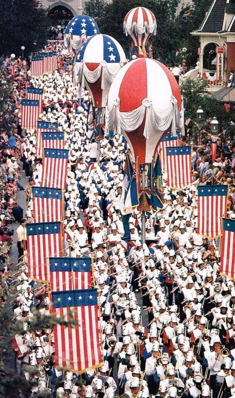 Marching band in white uniforms parades down a street, holding American flag banners. Large red, white, and blue hot air balloon floats are overhead. Crowds line both sides of the street. Patriotic decorations are everywhere.