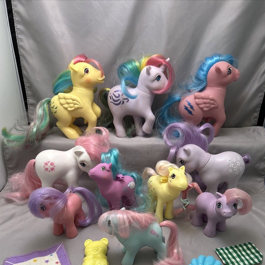 A group of colorful My Little Pony toys with pastel manes and tails are displayed on a gray fabric background. They have various symbols on their sides and are arranged in two rows, facing different directions.