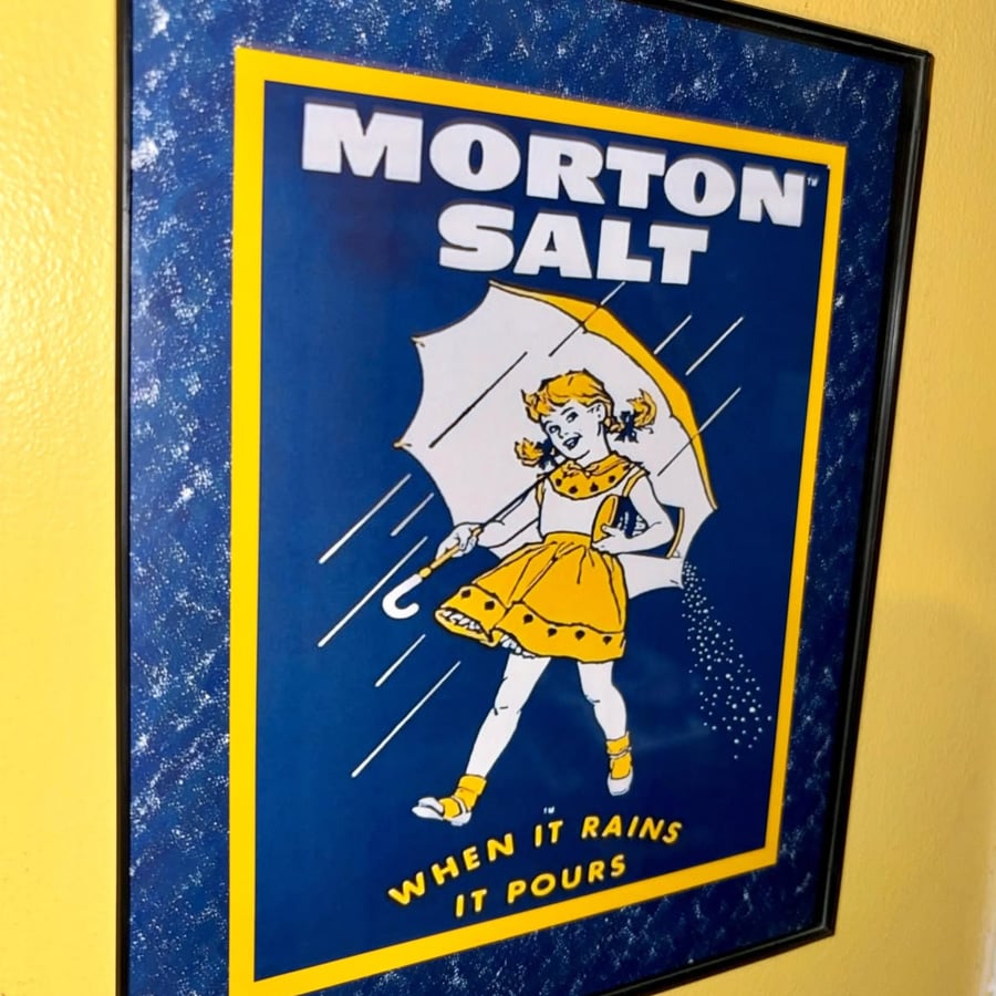 Framed Morton Salt poster shows a girl in a yellow dress holding an umbrella in the rain, with the text: "MORTON SALT - WHEN IT RAINS IT POURS.