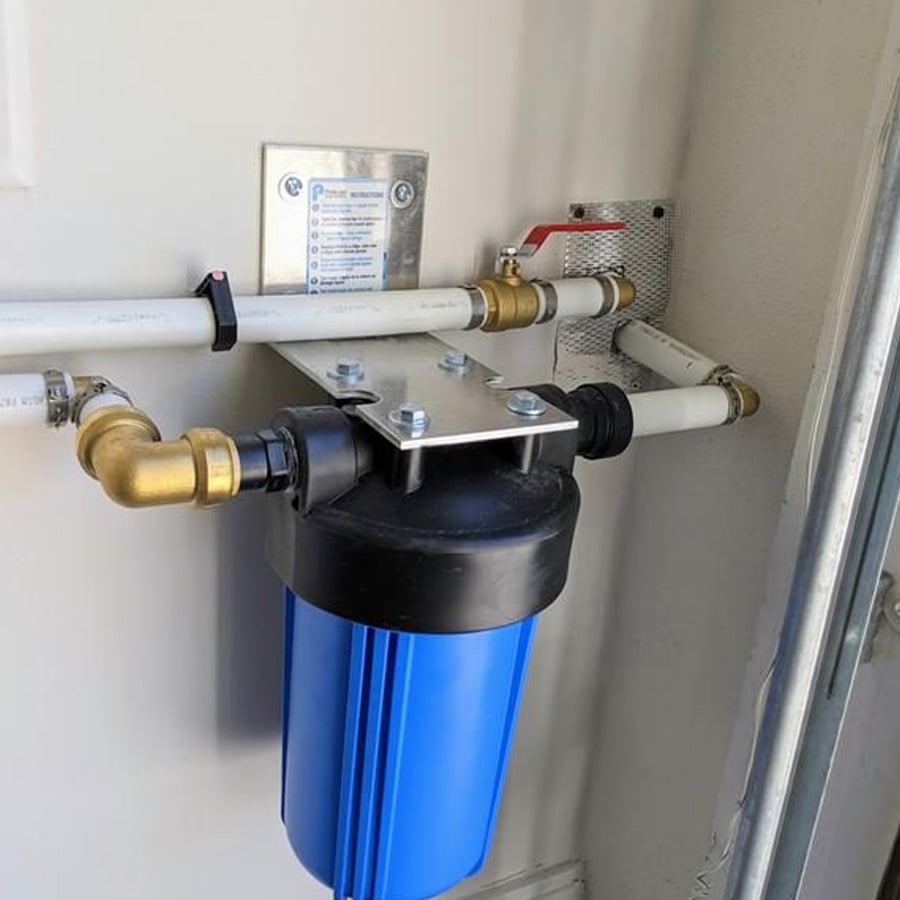 A blue water filter housing is mounted on a wall, connected to white PVC pipes with brass fittings and a red-handled valve. A small instruction label is posted above the filter.