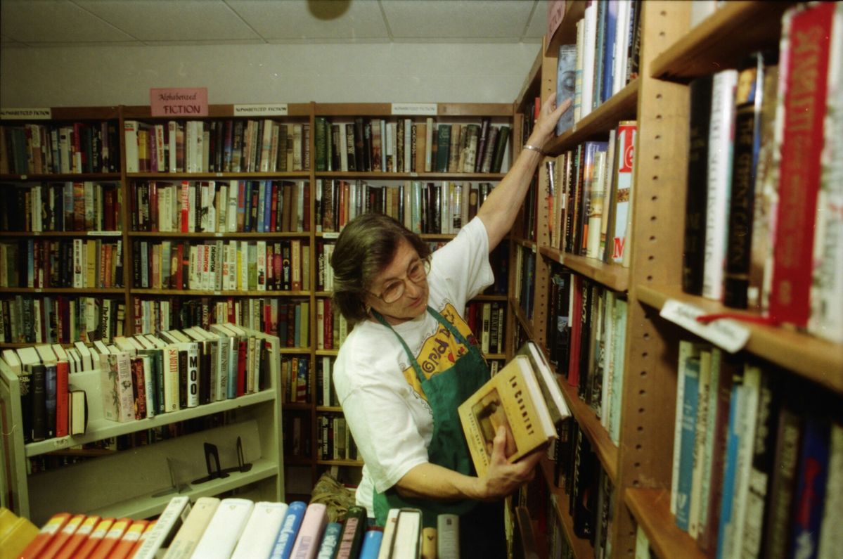 A person wearing glasses and an apron arranges books on a crowded bookstore shelf, reaching up with one hand and holding multiple books in the other. Shelves around them are filled with assorted books.