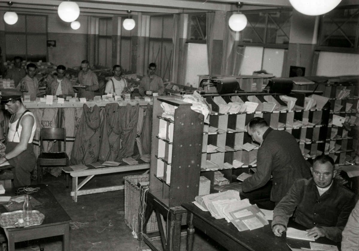 Black-and-white photo of workers sorting mail in a busy room with cubbyholes filled with envelopes, sacks of mail hanging behind, and papers stacked on desks. Several men are seated or standing, organizing and processing letters.