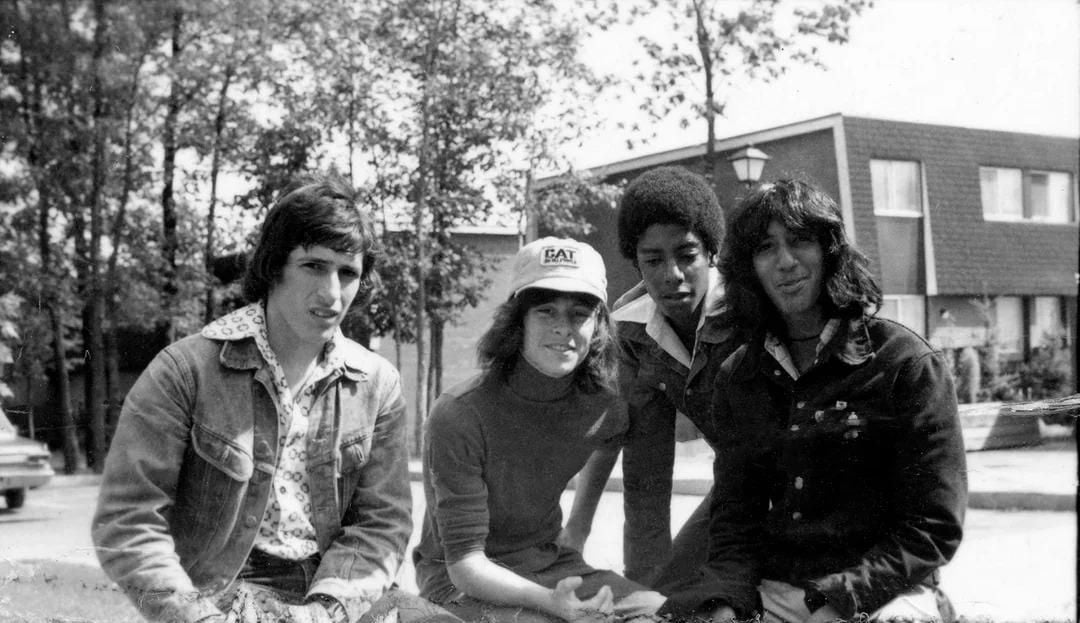 Four young people sit outdoors in front of houses and trees on a sunny day. Three are men, one is a woman wearing a baseball cap. They are smiling and dressed in casual 1970s-style clothing.