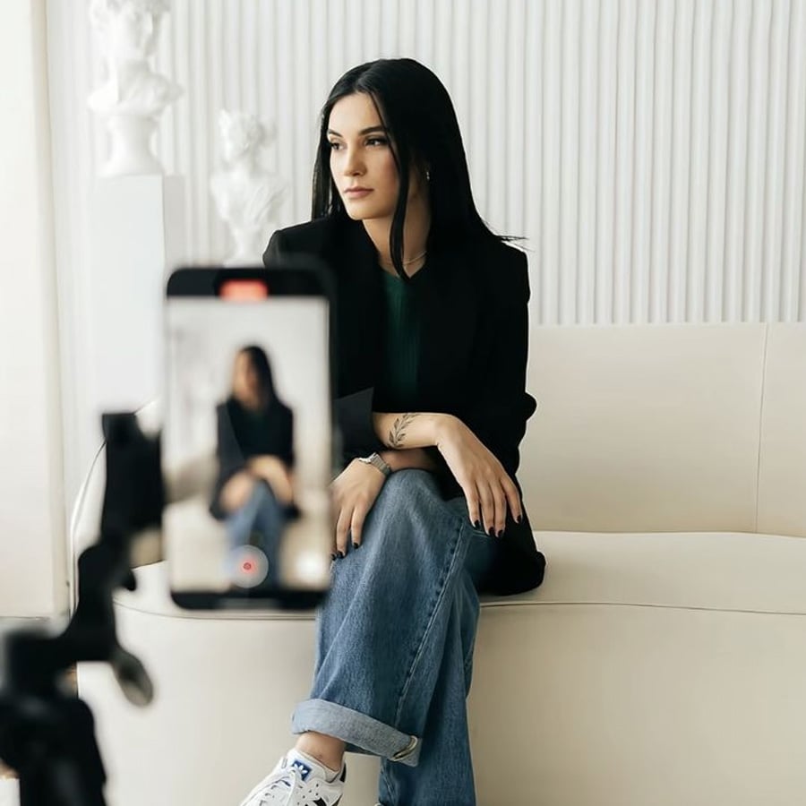 A woman with long dark hair sits on a cream sofa, posing for a video being recorded on a smartphone mounted on a tripod in a bright, minimal room.