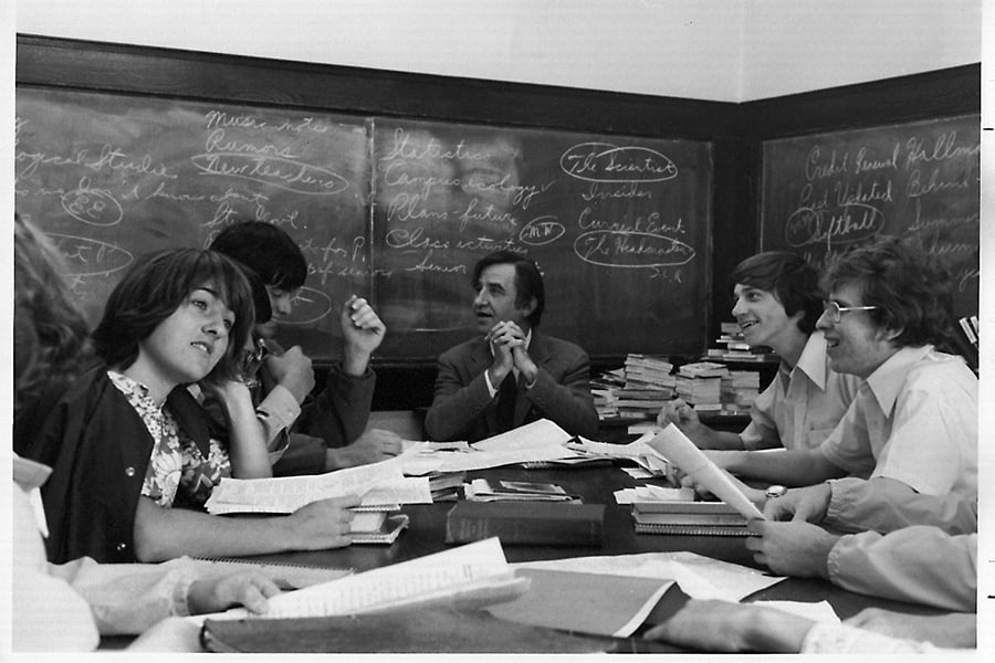 A group of students and a teacher sit around a table, engaged in discussion with books and papers. Chalkboard behind them is filled with writing and diagrams.