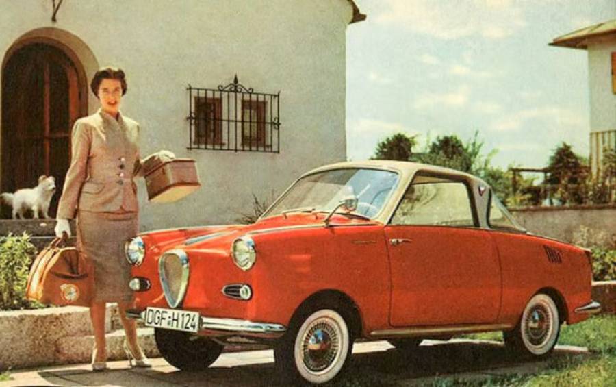 A woman in a suit stands beside a small red vintage car, holding luggage, with a white dog and a house in the background.