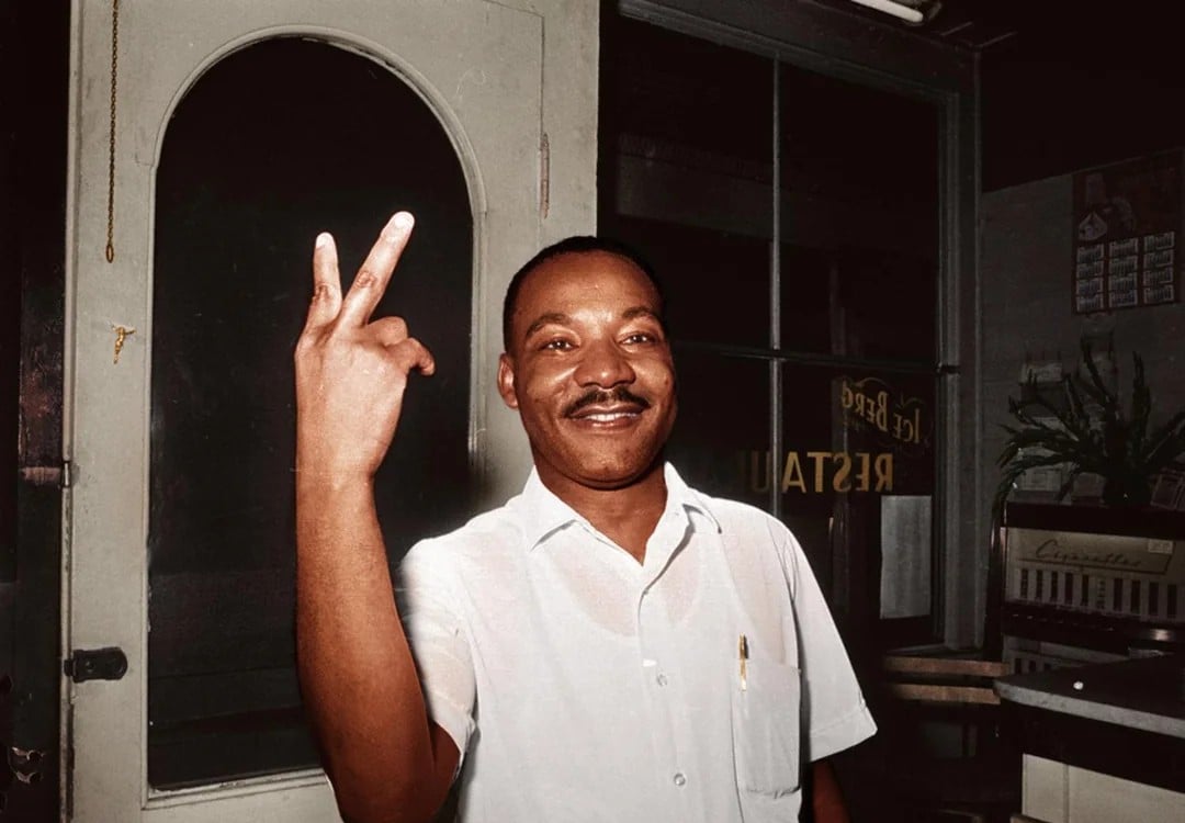 A man in a white shirt smiles and holds up two fingers in a peace sign, standing indoors near a door with glass panes.