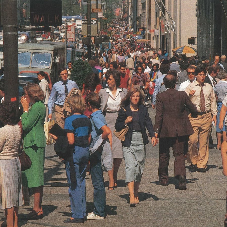 A busy city sidewalk filled with people walking in both directions on a sunny day. Many wear business attire, and buildings, vehicles, and street signs are visible in the background.
