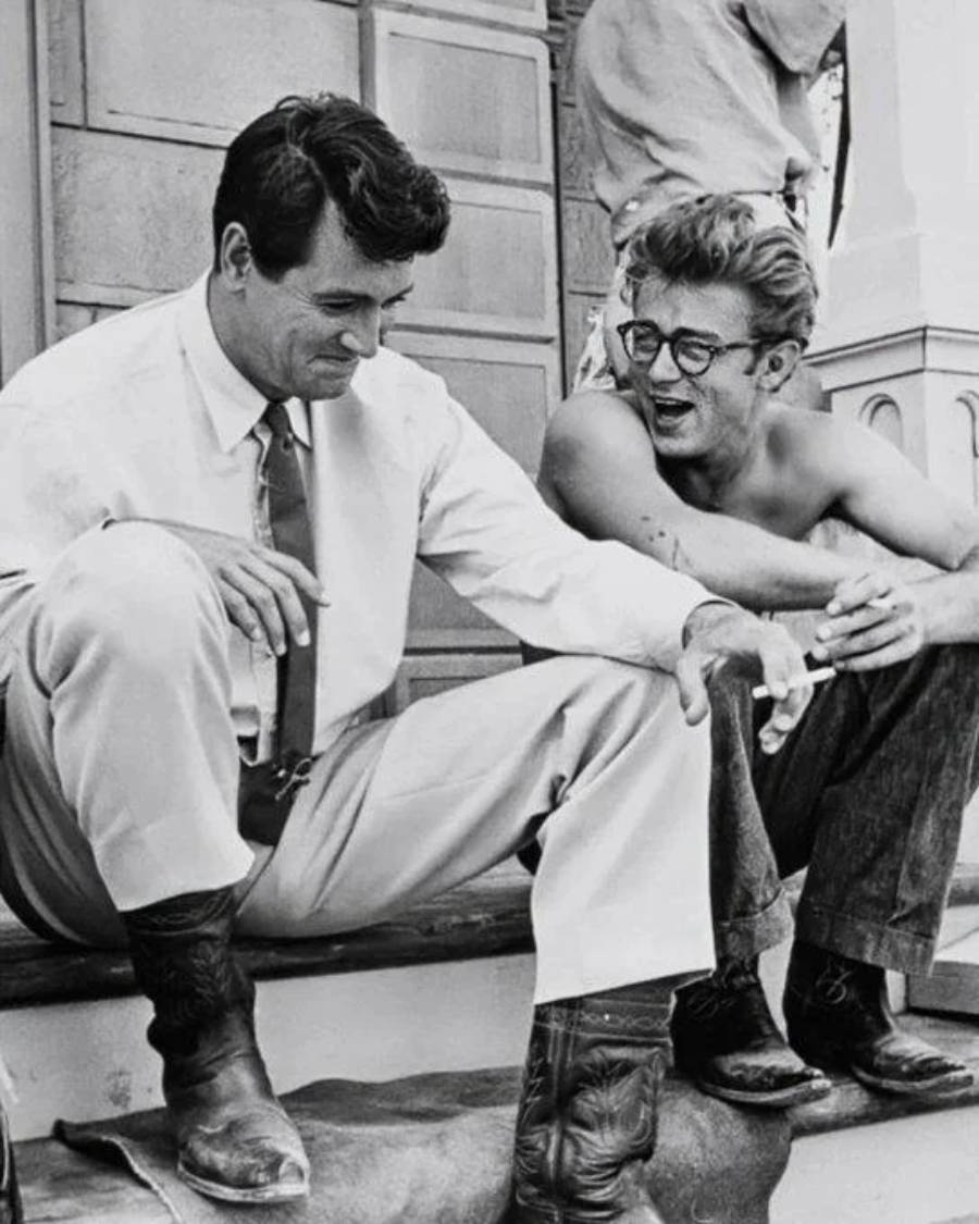 Two men sit on outdoor steps, smiling and talking. One man wears a shirt, tie, and cowboy boots, looking down. The other, shirtless with glasses, leans in, holding a cigarette, engaged in the conversation.