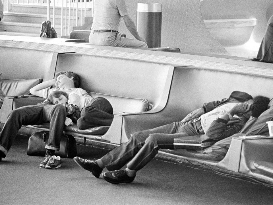 Three people are sleeping on curved seats in an airport waiting area, with their heads resting on their hands or arms. One person in the background sits upright, facing away. The scene appears calm and quiet.