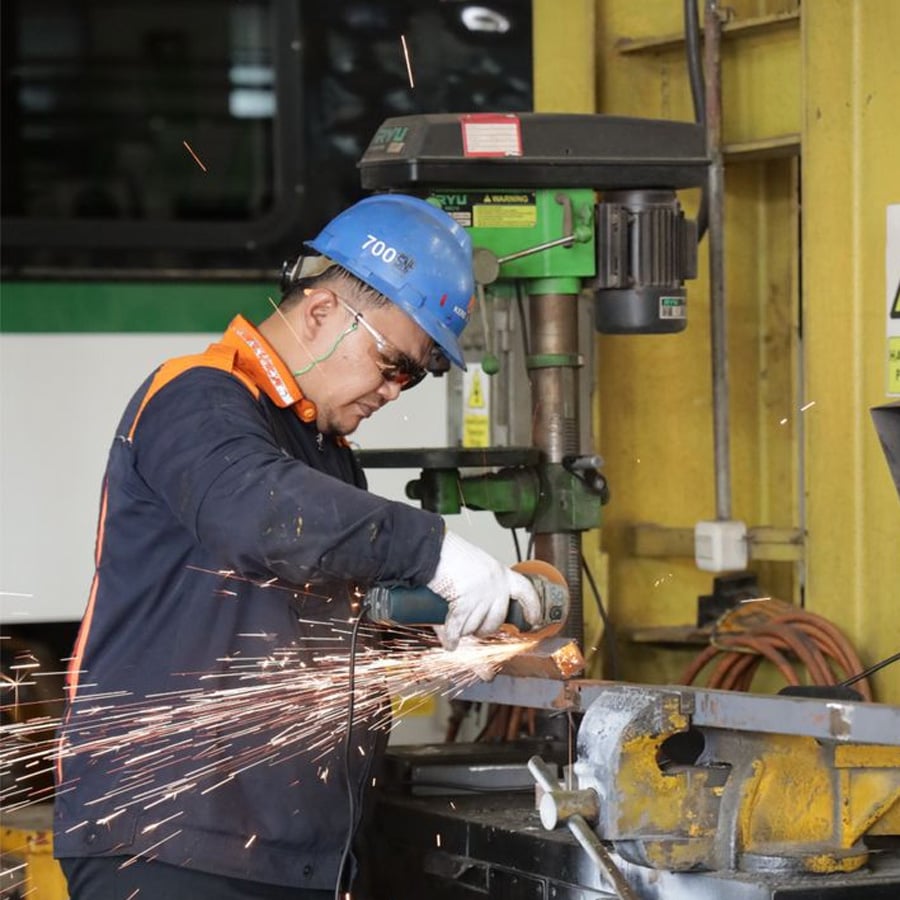 A worker in safety gear uses a grinder to cut metal, causing sparks to fly in a workshop with yellow walls and equipment in the background.