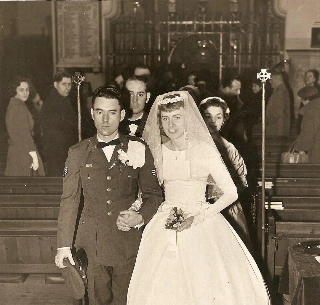 A bride and groom pose in a church. The groom wears a military uniform and holds his hat; the bride is in a white dress and veil, holding flowers. People stand and sit in pews behind them.
