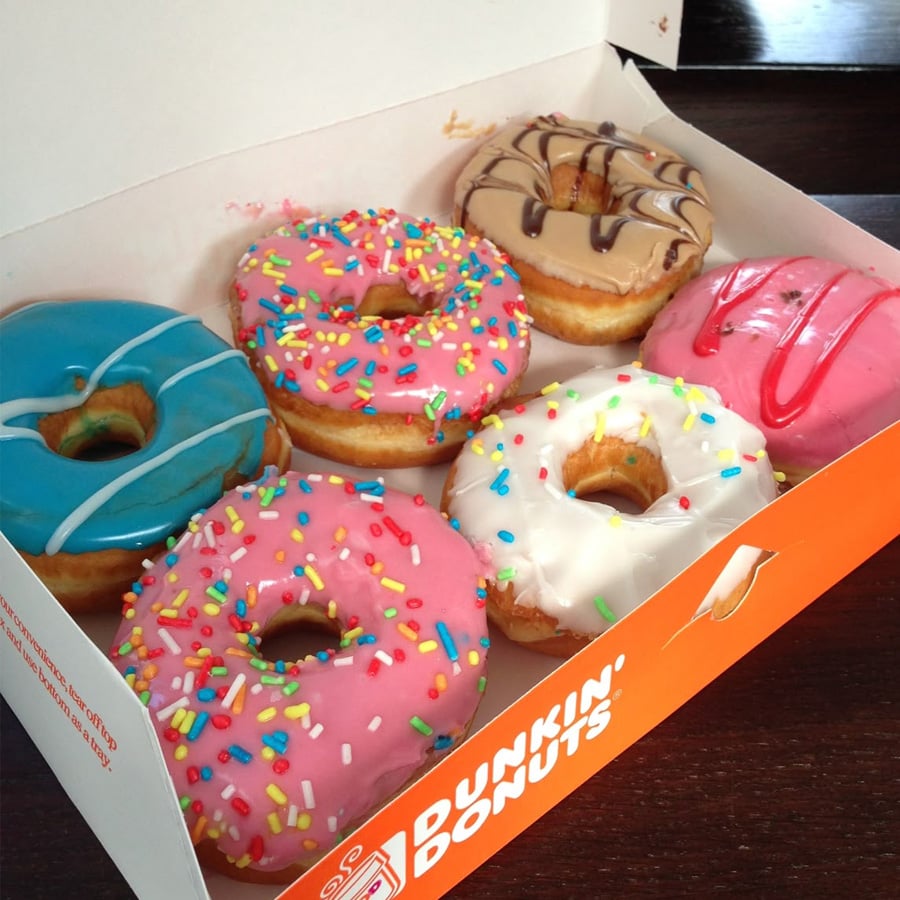 Six colorful donuts with various icing and sprinkles are arranged in an open Dunkin’ Donuts box on a dark table. The donuts are decorated in blue, pink, white, tan, and chocolate icing.