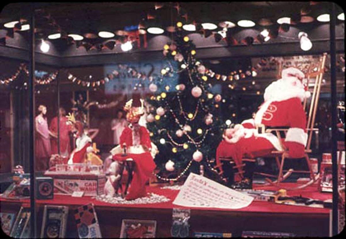 A festive store window display shows Santa in a rocking chair beside a decorated Christmas tree, surrounded by gifts, toys, and holiday decorations.