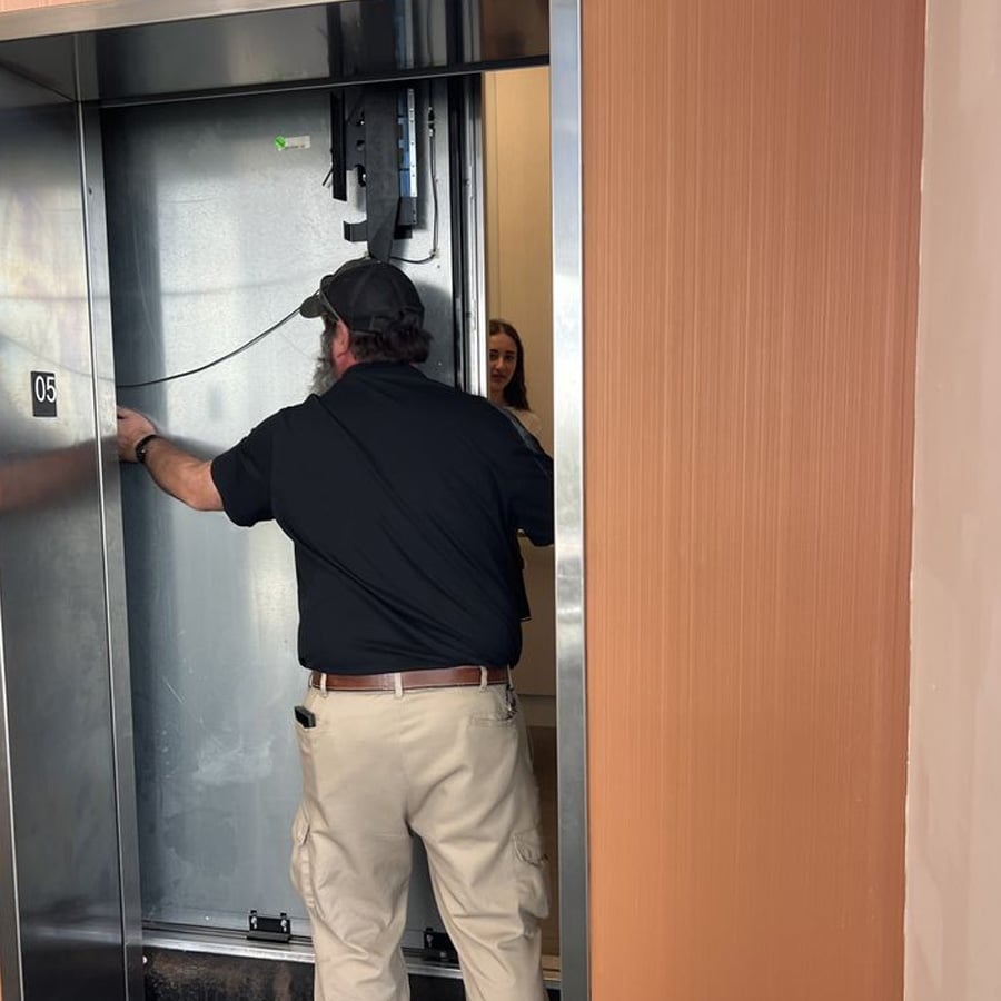 A man in a black shirt and khaki pants is repairing an elevator while a woman stands inside, visible through the partially open elevator doors.