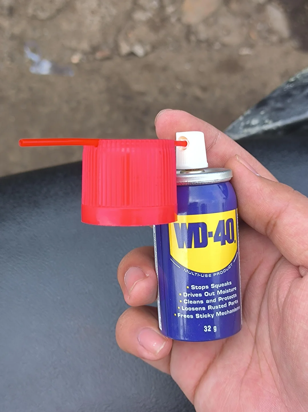 A hand holds a small 32g can of WD-40 with a red spray nozzle attached. The background is blurred with some dirt and a black surface visible.