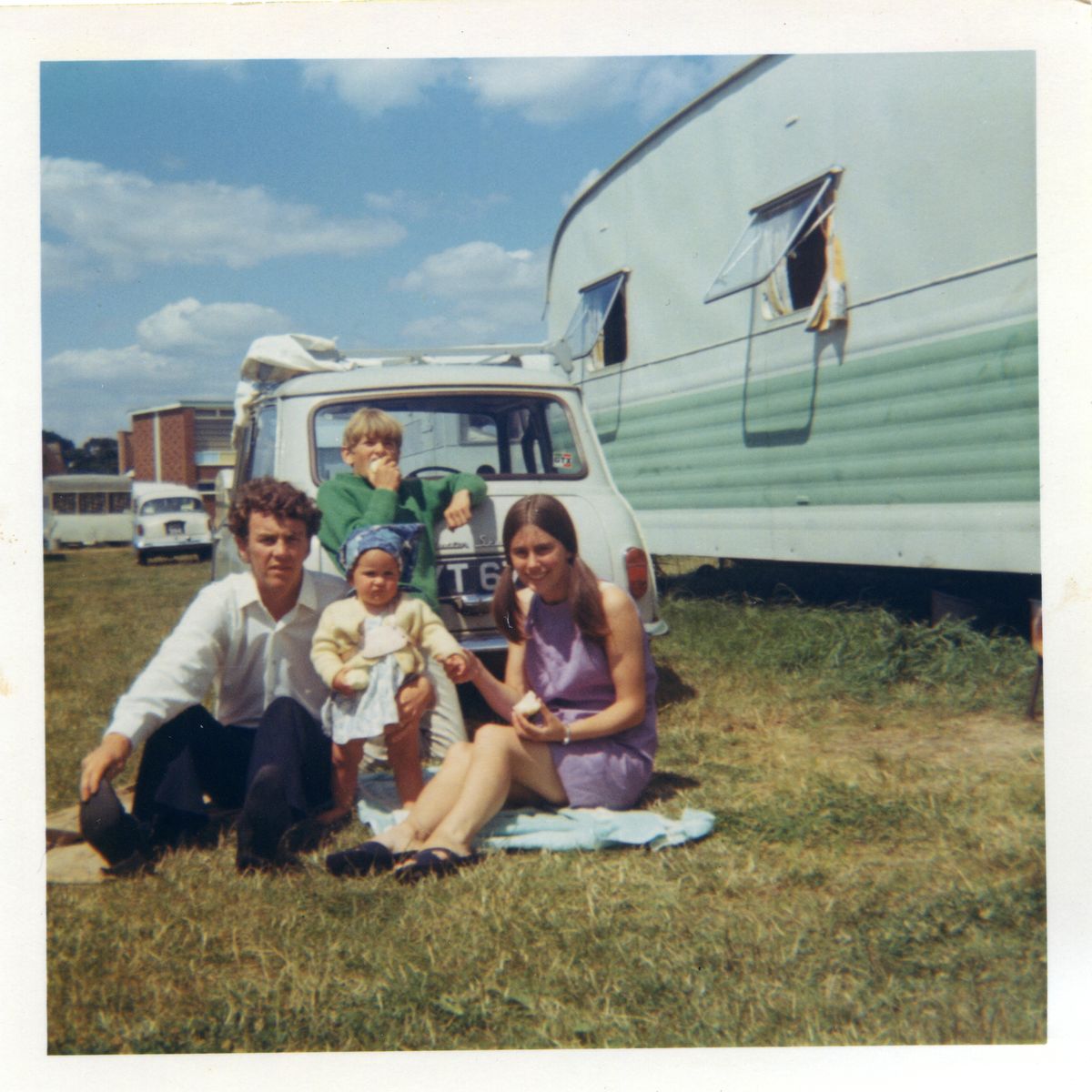A family of four sits on a blanket beside a white car and a green trailer on a grassy field, enjoying a sunny day. The parents sit with their two young children.