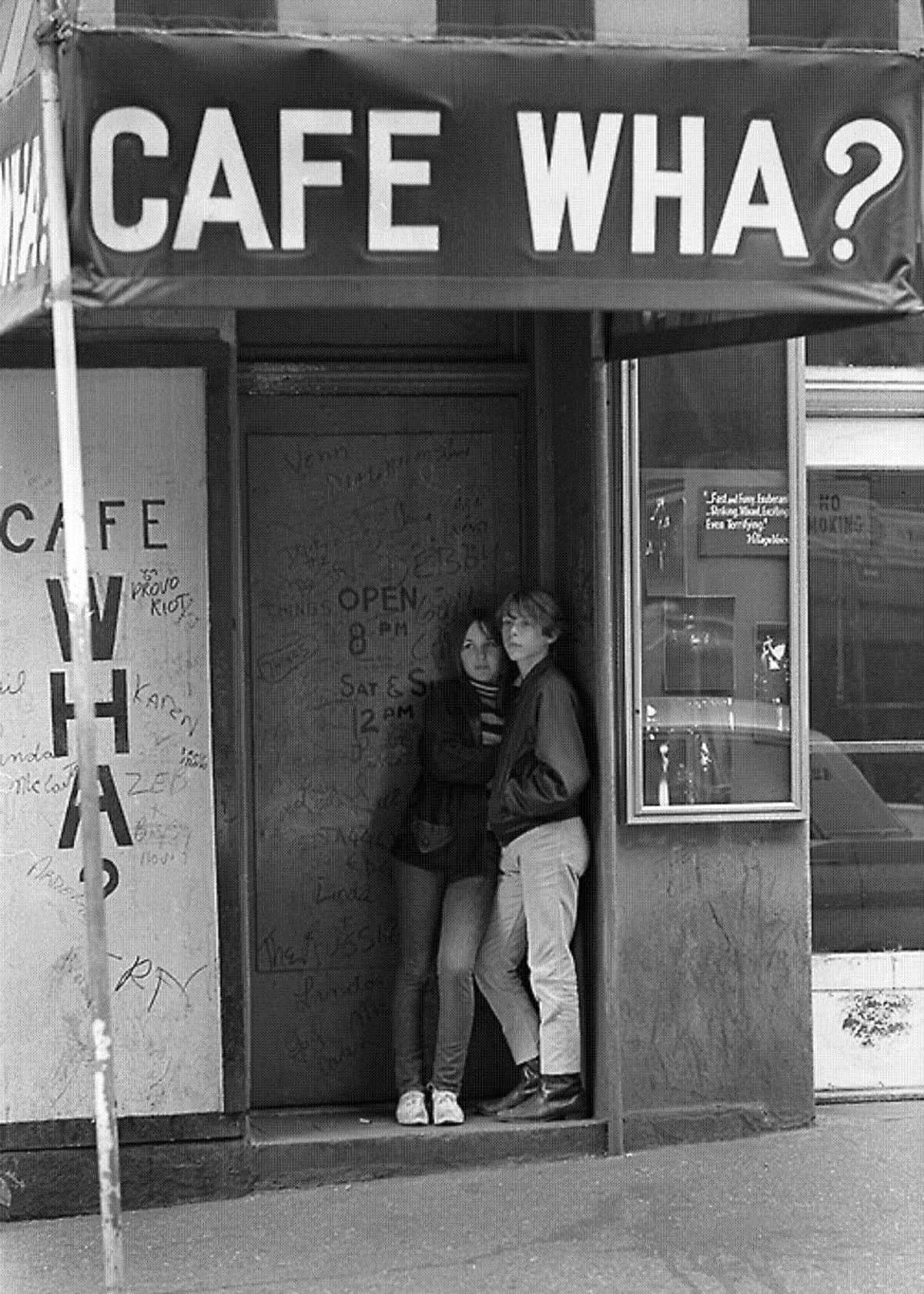 Two people stand closely together in the doorway of Cafe Wha?, a club with graffiti on the door and a sidewalk sign above them. The scene is in black and white, creating a nostalgic atmosphere.
