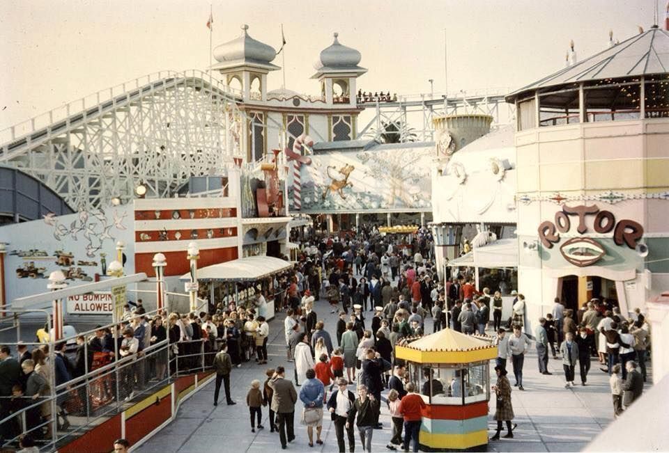 A crowded amusement park scene with people walking among colorful booths, a large wooden roller coaster, carnival games, and a ride called "ROTOR" with a painted face on its entrance.