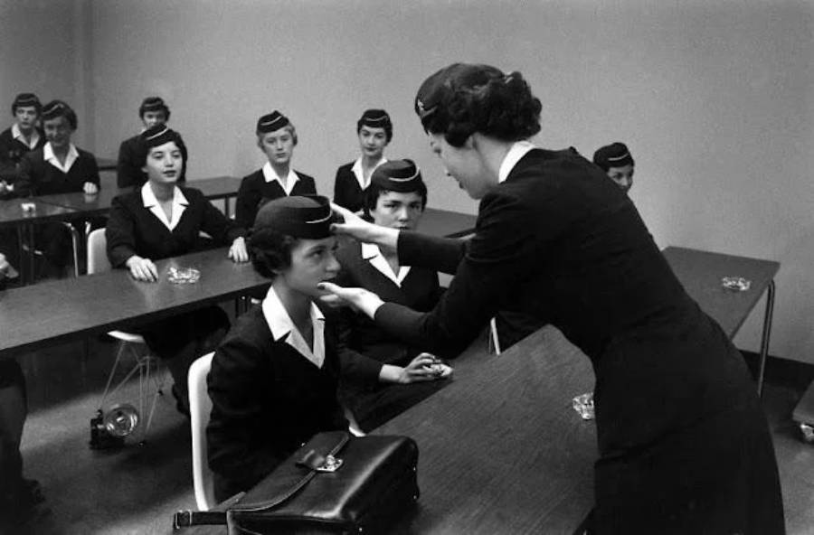 A woman in uniform adjusts another woman’s hat in a classroom, while several other women in matching uniforms sit at desks and watch attentively. The scene appears to be a training session from the mid-20th century.