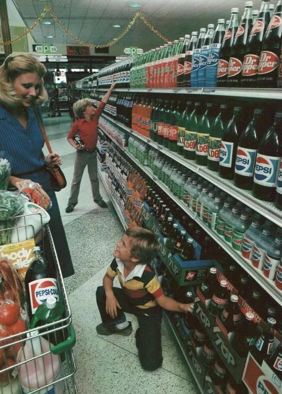 A woman with a shopping cart full of groceries stands in a supermarket aisle as two young boys excitedly reach for soda bottles on the shelves, with brands like 7UP, Pepsi, and Dr Pepper visible.