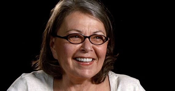 A middle-aged woman with short brown hair and glasses smiles while looking slightly off camera. She is wearing a light-colored top and is set against a plain black background.
