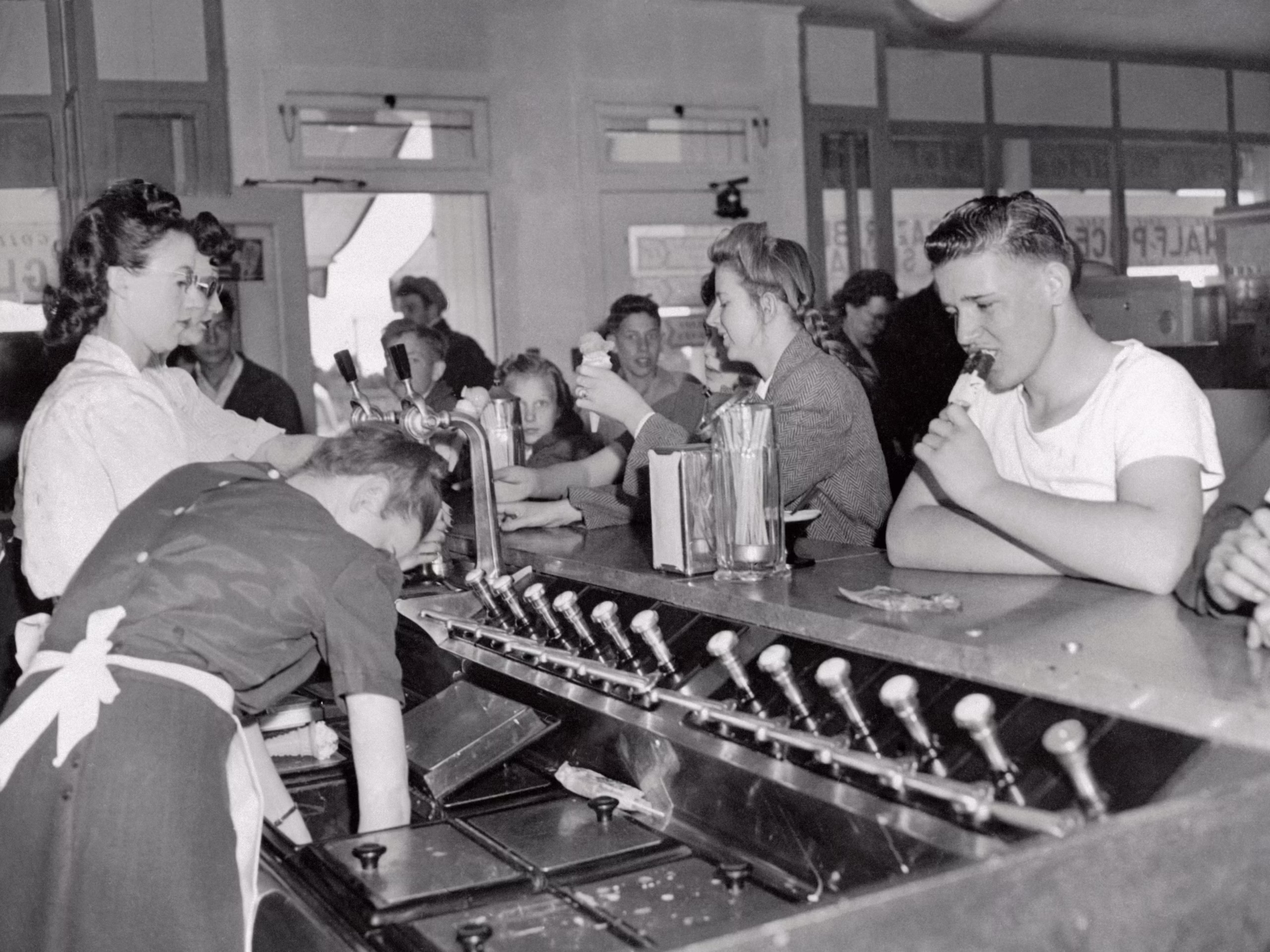 Black-and-white photo of a busy ice cream parlor, with a server and customers at the counter. A boy eats an ice cream cone while others order or chat in the background. The scene is lively and nostalgic.