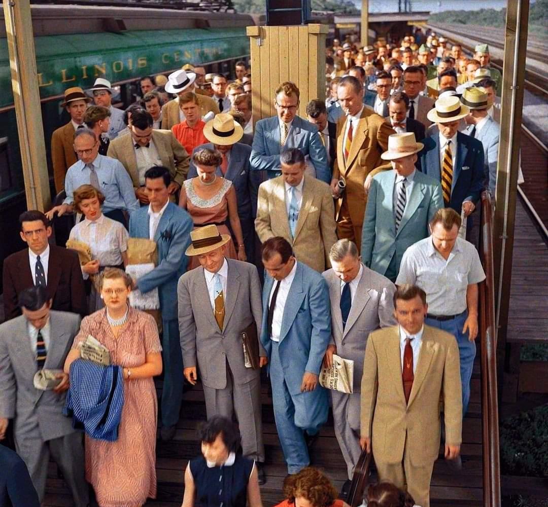 A large group of people in 1950s attire, many in suits and hats, walk down a train station platform. A green train is visible on the left. The scene appears busy and lively.