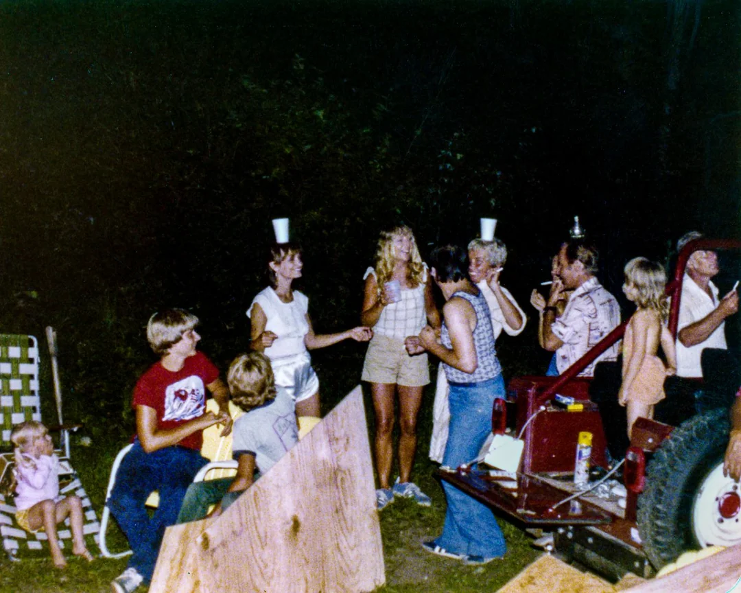A group of people at an outdoor gathering at night; some balance cups on their heads while others watch or chat. A red vehicle is nearby, and children sit on lawn chairs.