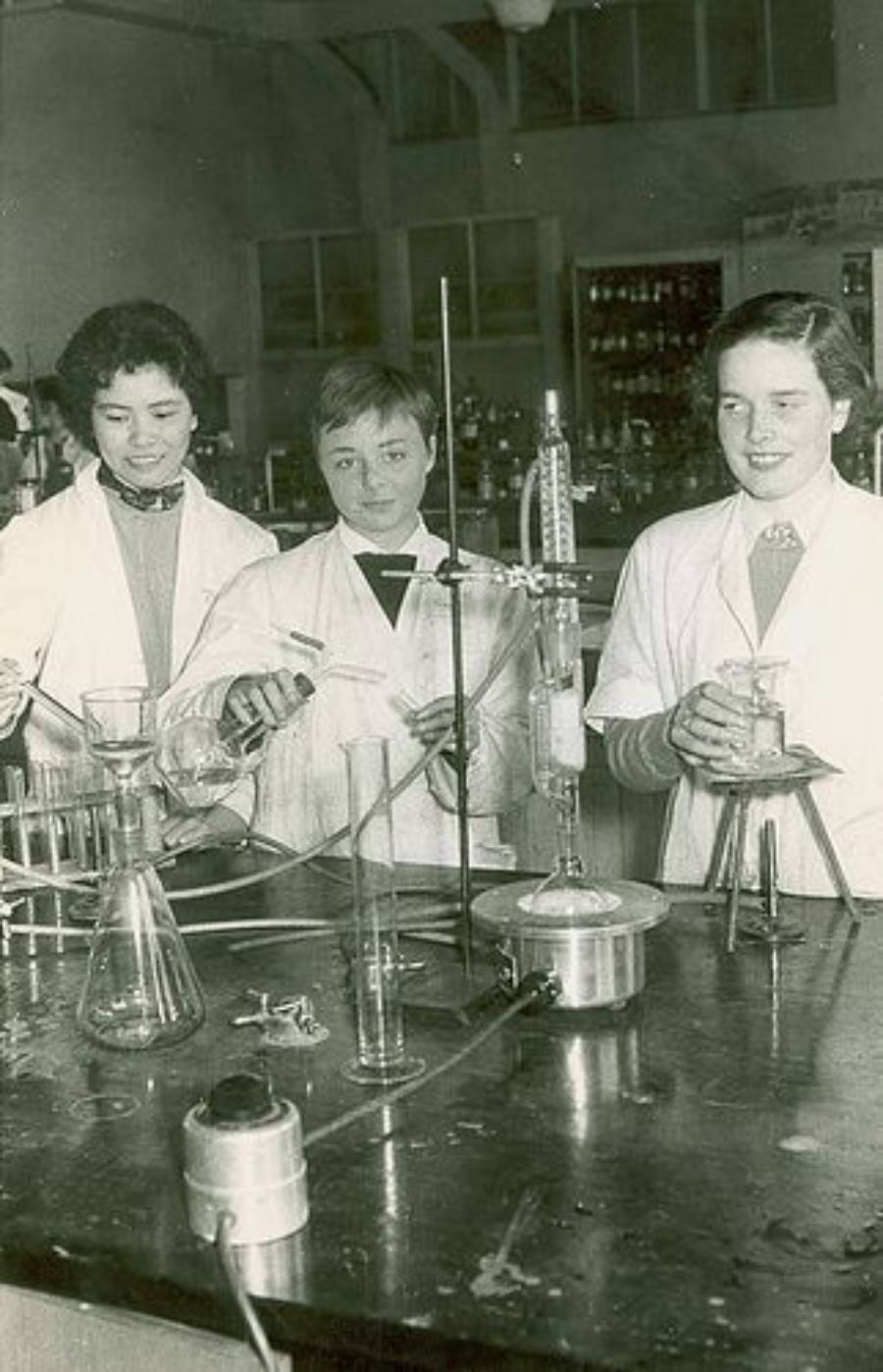 Three women in lab coats stand at a workbench in a science laboratory, working with glassware and scientific equipment. Shelves with bottles and lab items are visible in the background.