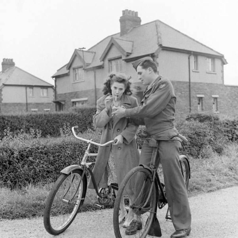 A woman and a man in uniform sit on bicycles by a hedge. The man lights the woman’s cigarette. Two houses are visible in the background on a cloudy day.