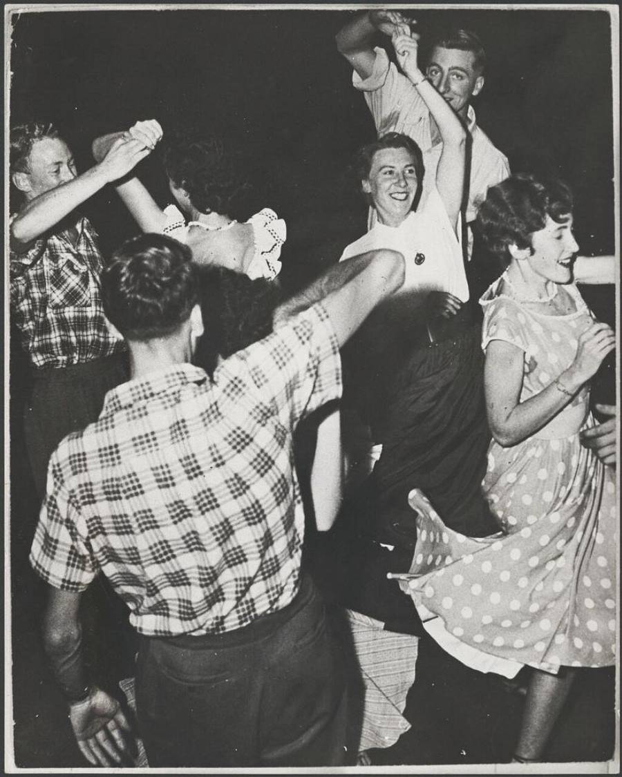 A group of young people, dressed in 1950s clothing, are joyfully dancing together. Some are holding hands and twirling, and everyone looks happy and energetic. The scene is lively and captures a festive moment.