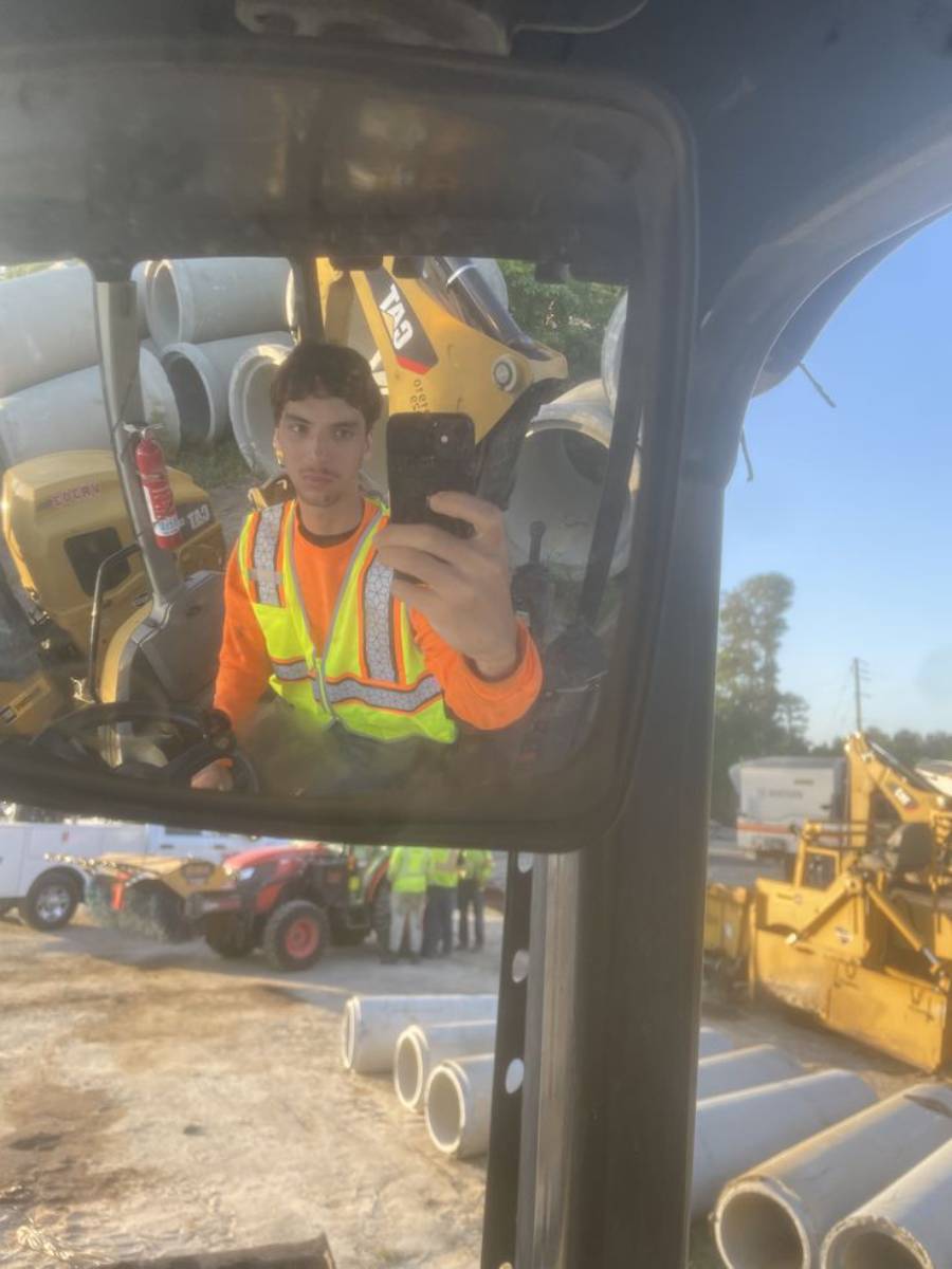 A man in a neon safety vest and orange shirt takes a selfie in a construction vehicle mirror at a worksite with pipes, equipment, and crew visible in the background.