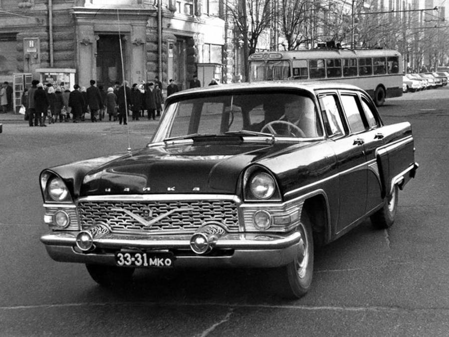 A classic black sedan with chrome details drives on a city street; a group of people and a trolleybus are seen in the background, with buildings and leafless trees lining the street.