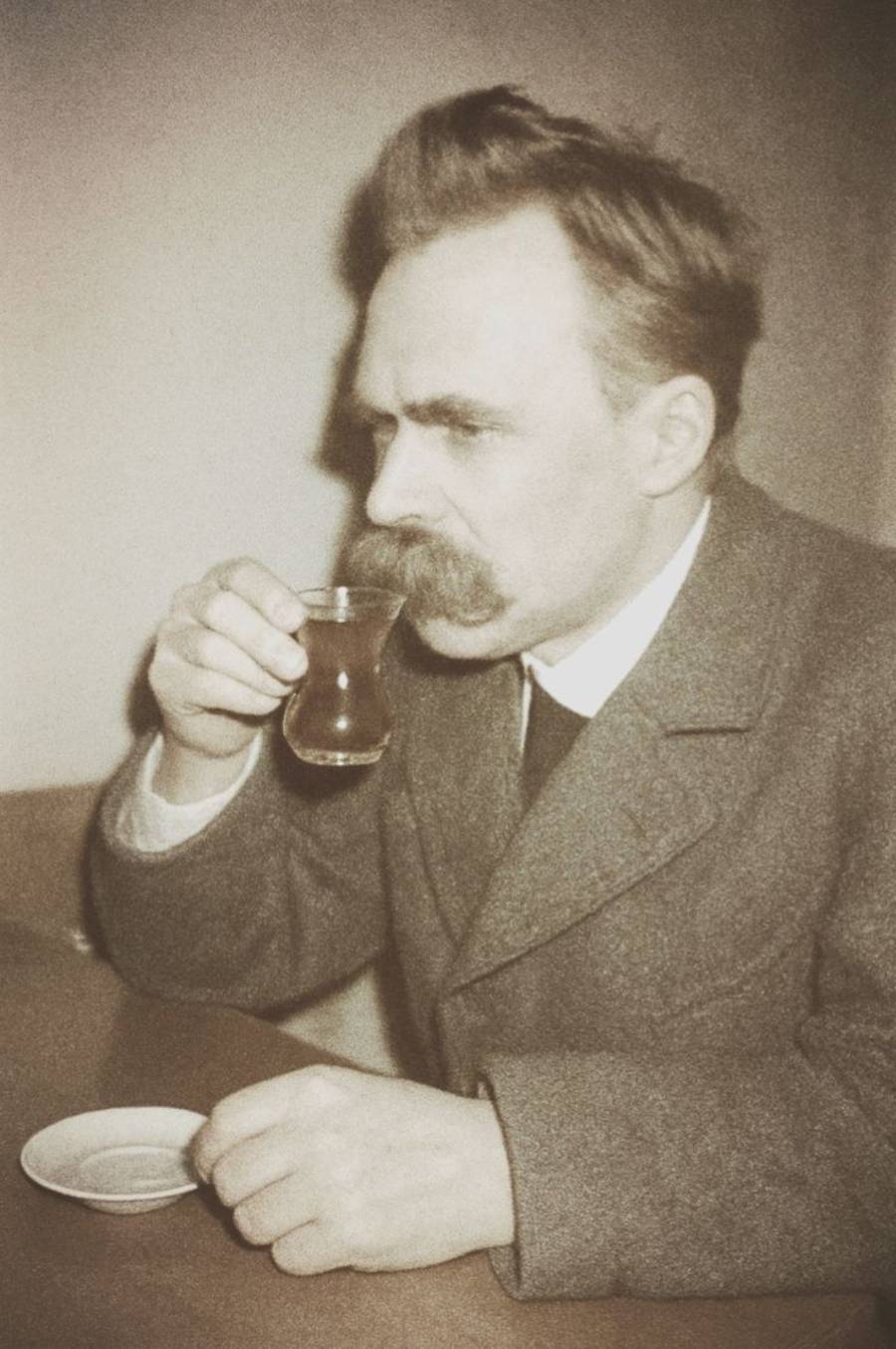 A man with a thick mustache and styled hair sits at a table, holding a glass of tea close to his face. He wears a suit jacket and looks thoughtful. A small saucer is on the table in front of him.