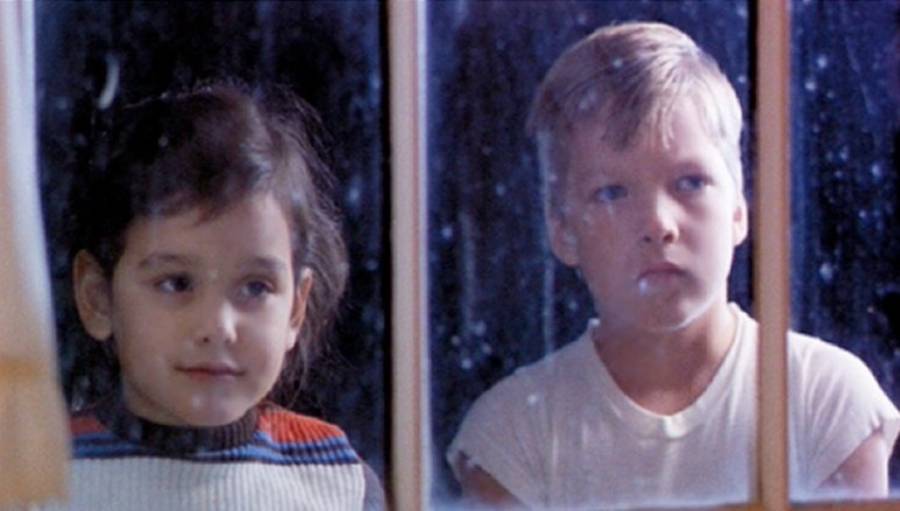 Two young children, a girl and a boy, look outside through a window at night. The girl is smiling softly, while the boy has a neutral expression. Reflections and raindrops are visible on the glass.