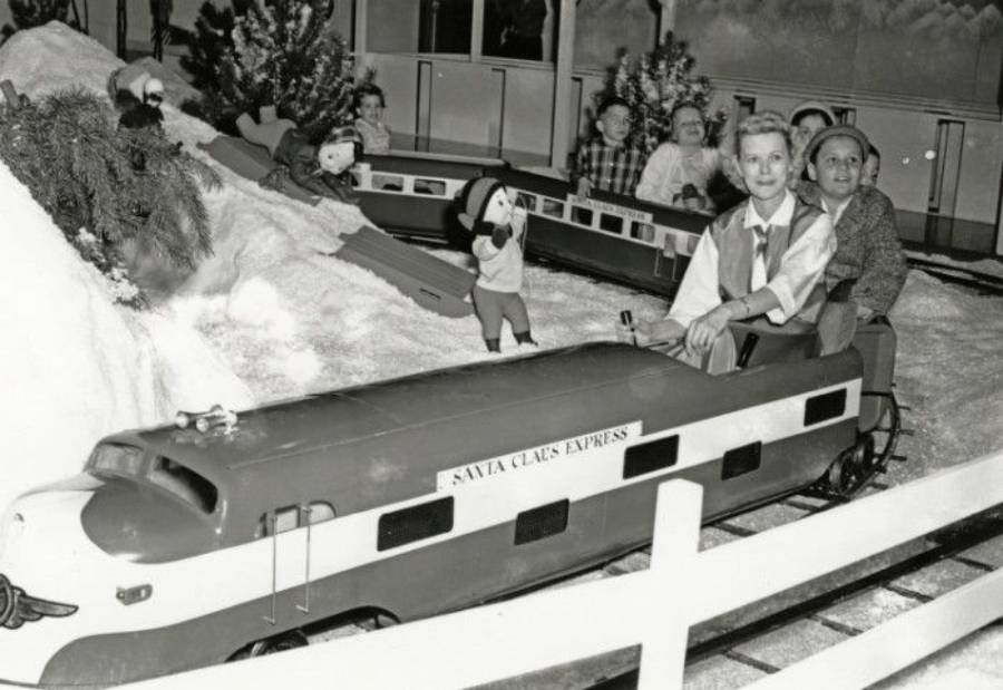 Children ride a small train labeled "Santa Claus Express" at a snowy indoor amusement setup, with festive decorations, trees, and dolls in the background.