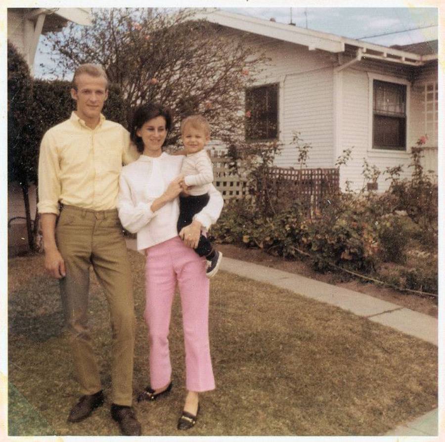A man, woman, and baby pose together in a yard in front of a white house with a garden and a wooden fence. The woman holds the baby, and all three smile at the camera. The photo has a vintage feel.
