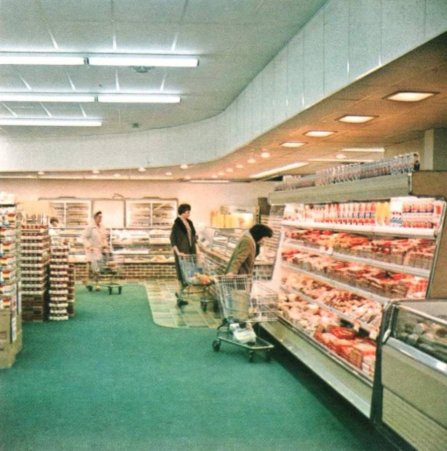 A supermarket aisle with green carpet, people pushing shopping carts, and shelves stocked with groceries, dairy, and packaged foods under bright fluorescent lighting.