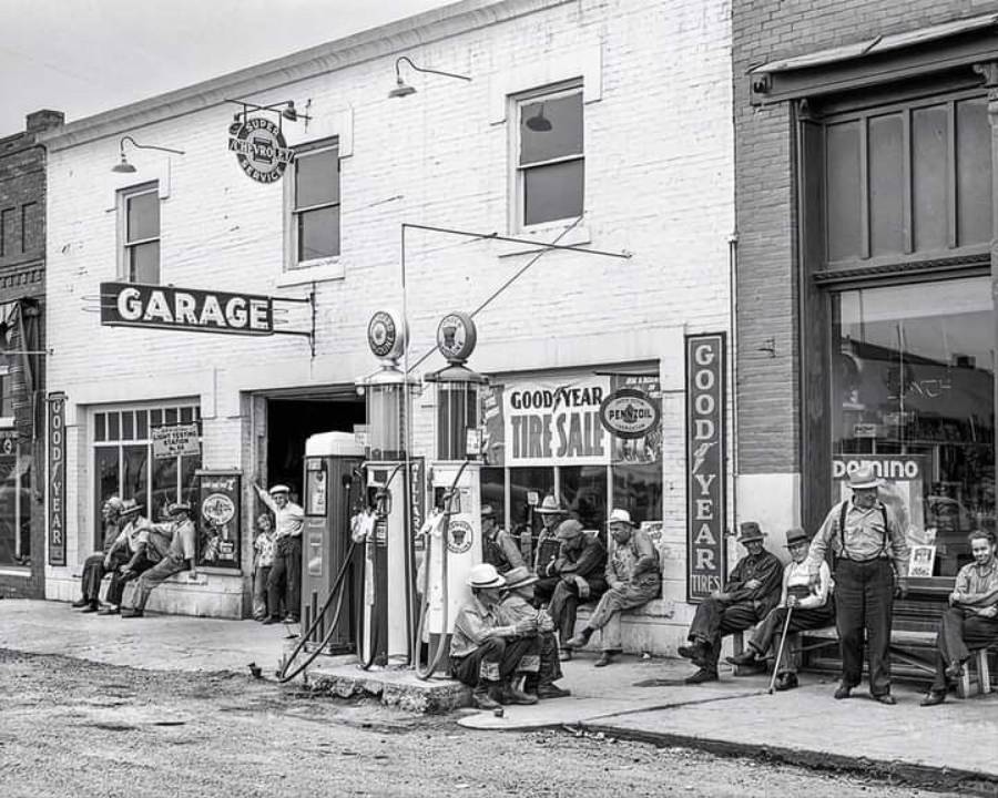 A group of men sit and stand outside a vintage garage and service station with old gas pumps, tire advertisements, and signs, likely from the early to mid-20th century. The scene has a small-town, nostalgic feel.
