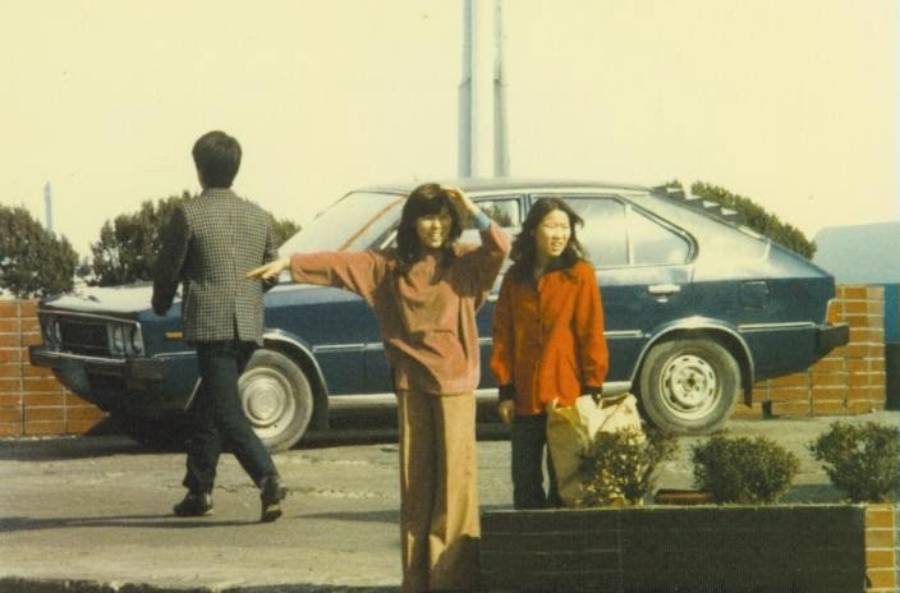 Three people stand near a parked blue car; one woman in brown waves her arm, another in red holds a bag, and a man walks away. There are low plants and a brick wall in the background.